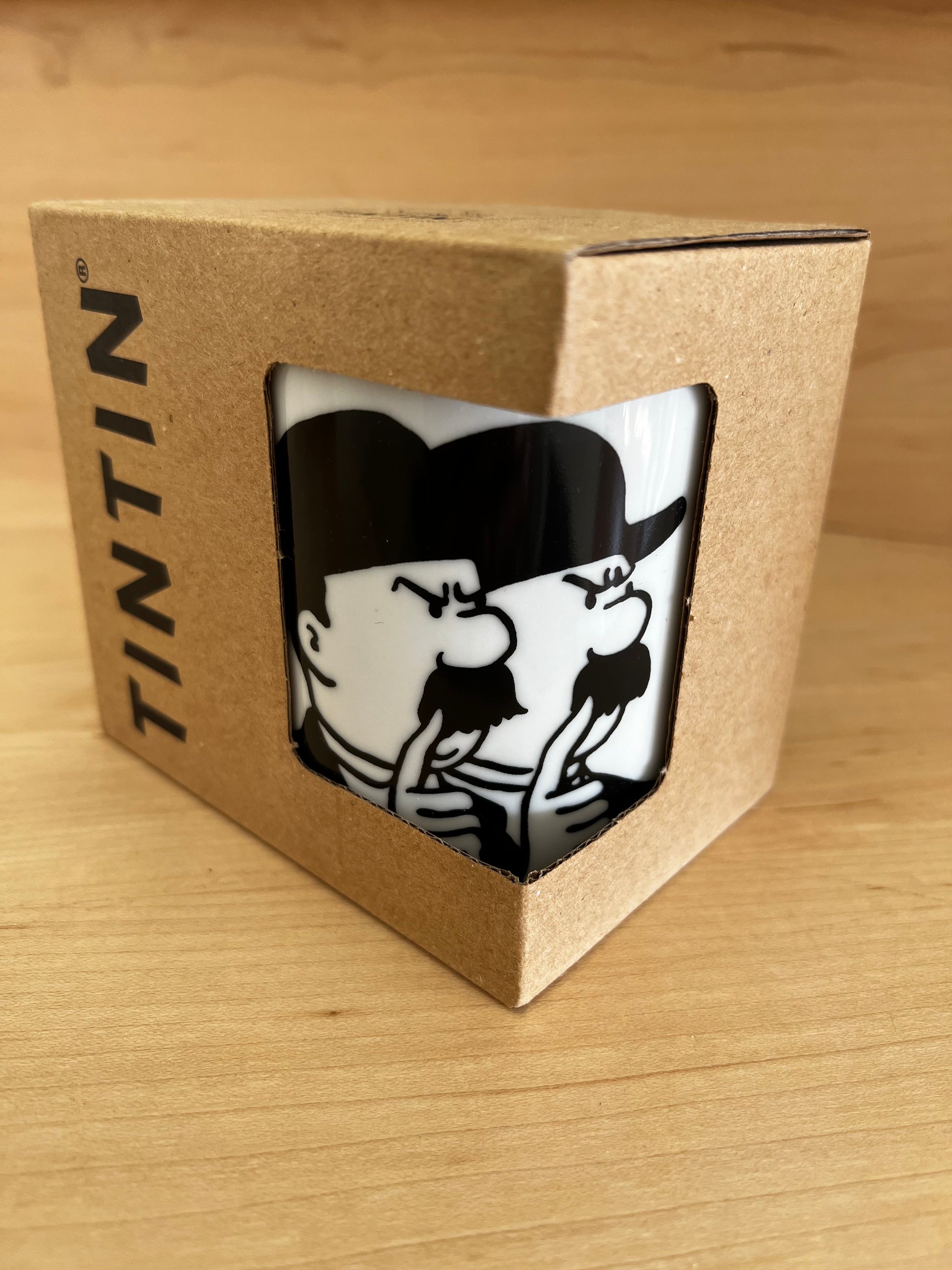 Close-up of a white porcelain Thomson and Thompson mug on a light wooden surface, featuring a bold, minimalist black line-art illustration of the two iconic bumbling detectives in their signature bowler hats and mustaches, holding their fingers to their lips in a secretive, whispering gesture.