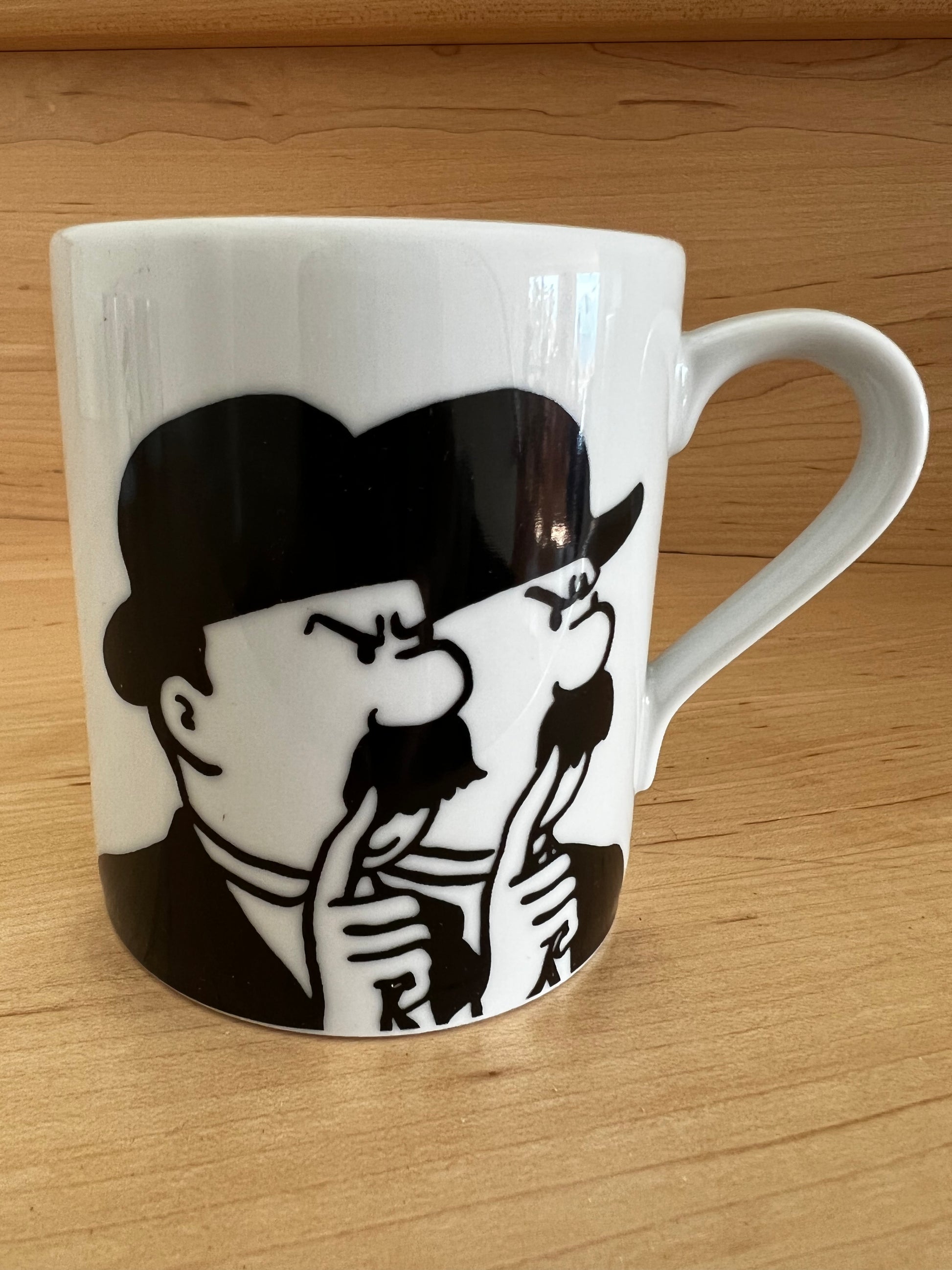 Close-up of a white porcelain Thomson and Thompson mug on a light wooden surface, featuring a bold, minimalist black line-art illustration of the two iconic bumbling detectives in their signature bowler hats and mustaches, holding their fingers to their lips in a secretive, whispering gesture.