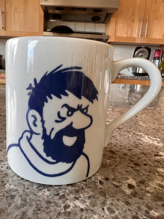 Close-up of a white porcelain Captain Haddock mug on a granite countertop in a kitchen setting, showing a bold, minimalist dark blue line-art illustration of Captain Haddock’s face, captured with a cheerful expression, matching his classic comic book style.