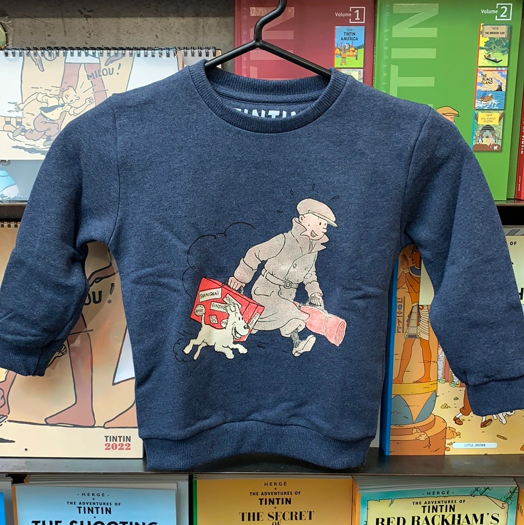 denim heathered kid's crewneck sweatshirt featuring classic Hergé comic art of Tintin in a flat cap sprinting alongside Snowy, who is carrying a bright red suitcase covered in Far East travel stickers, displayed on a hanger in front of Tintin book covers. 