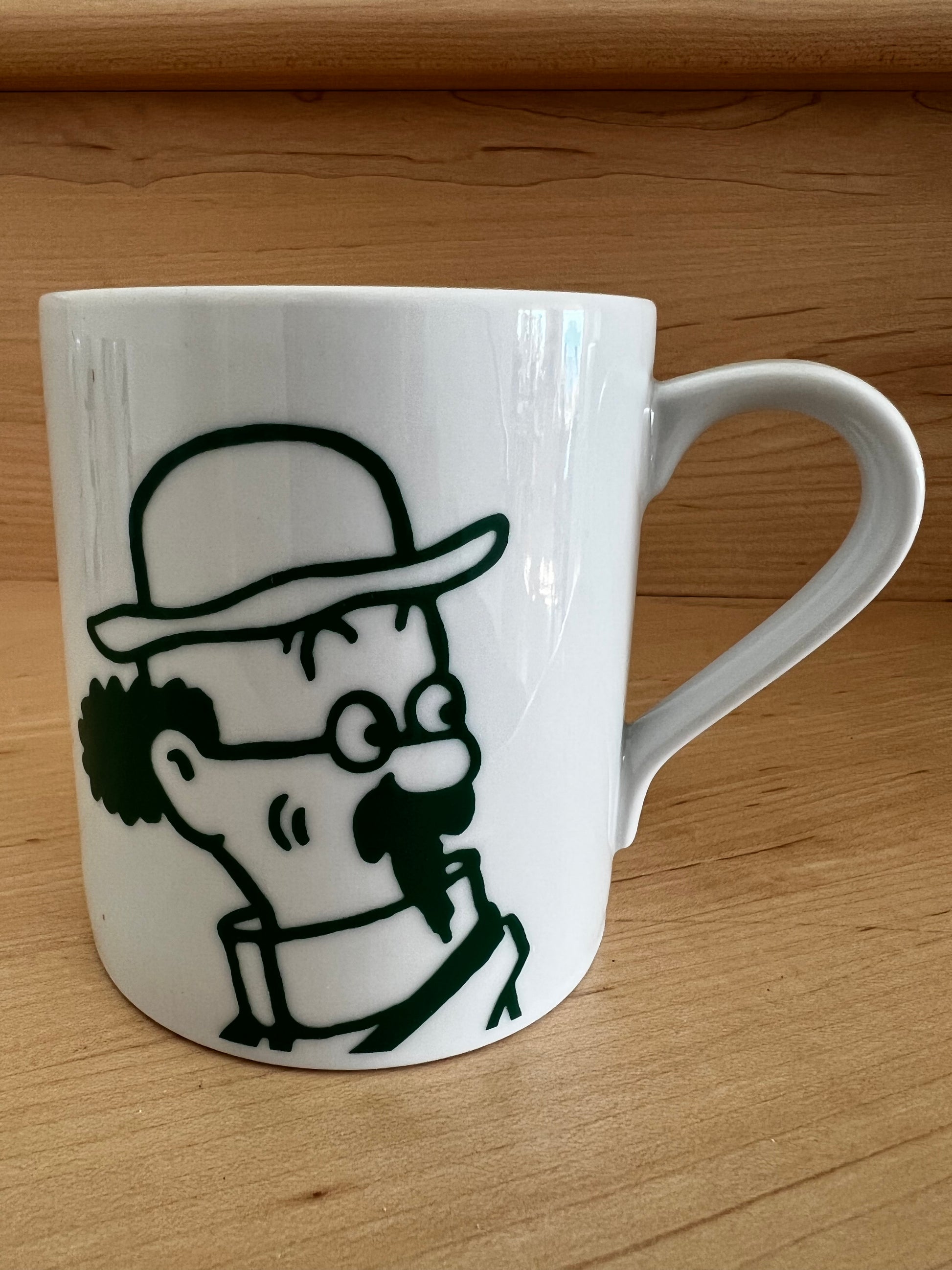 Close-up of a white porcelain Professor Calculus mug resting on a warm wooden surface, featuring a minimalist green line-art illustration of the brilliant, eccentric professor wearing his signature high collar and spectacles, captured in his classic thoughtful pose.