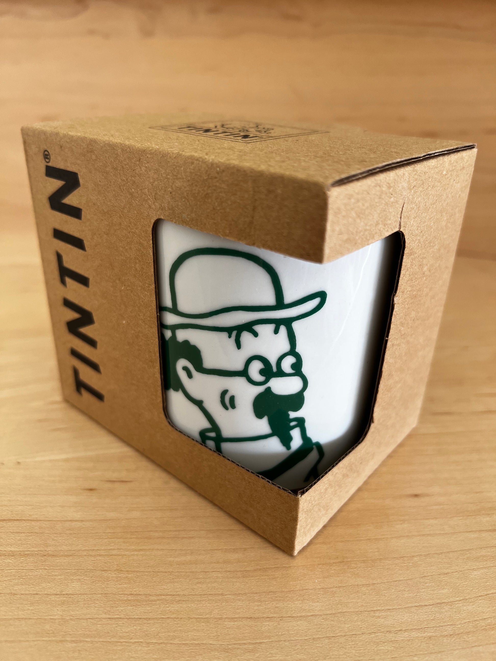 Angled front view of the white porcelain Professor Calculus mug packaged inside its open-front, kraft cardboard gift box with the black "TINTIN" logo printed vertically on the side. The mug inside displays the minimalist green line-art portrait of the eccentric professor, resting on a light wooden table.