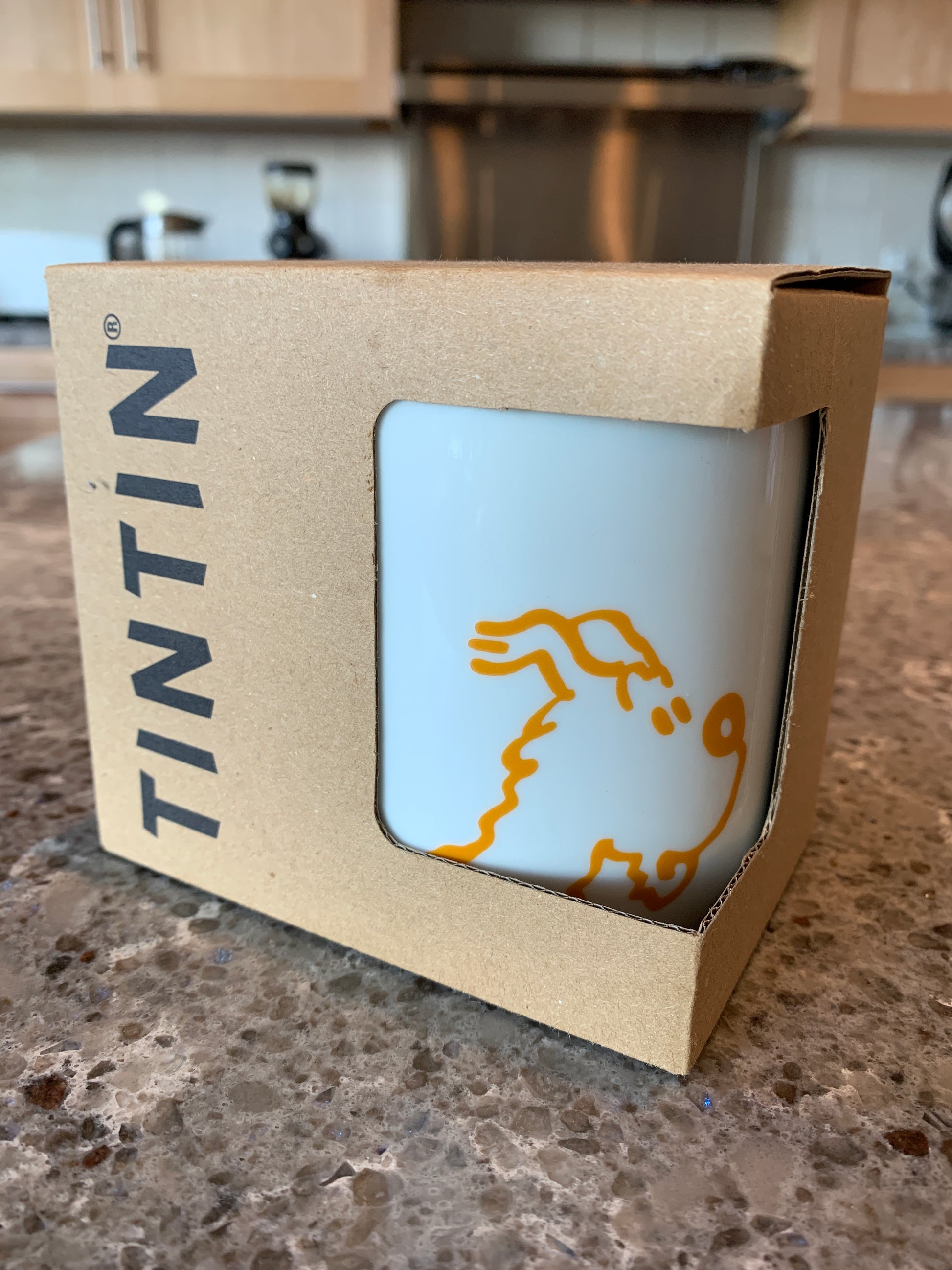 Angled front view of the white porcelain Surprised Snowy mug nestled inside its open-front, kraft paperboard gift box, featuring the black "TINTIN" logo printed vertically on the side. The mug displays the bold, minimalist orange line-art illustration of Snowy with his wide-eyed, startled expression.