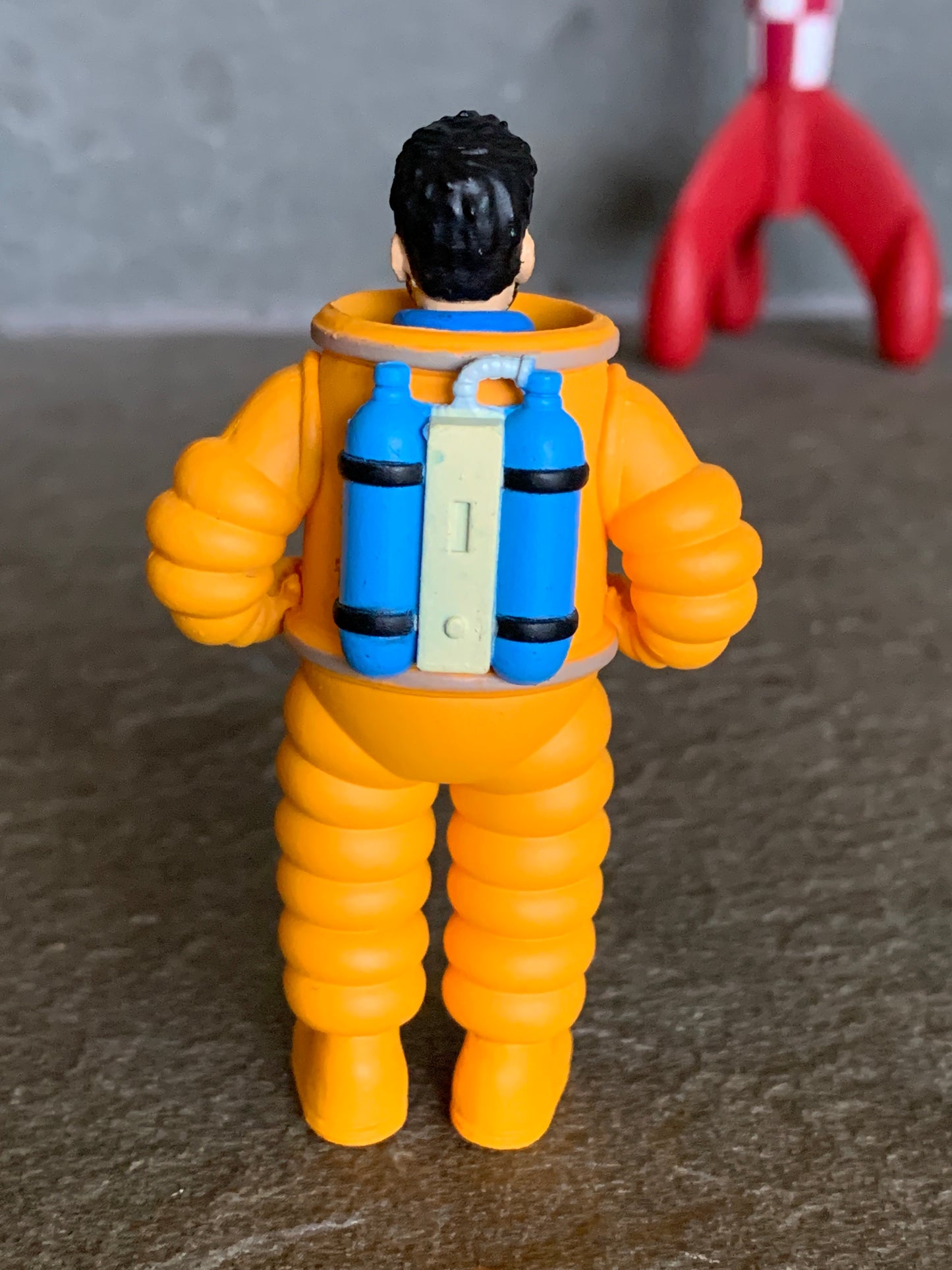 Captain Haddock cosmonaut mini figure in orange spacesuit with blue oxygen tanks, facing away from camera, red rocket in background