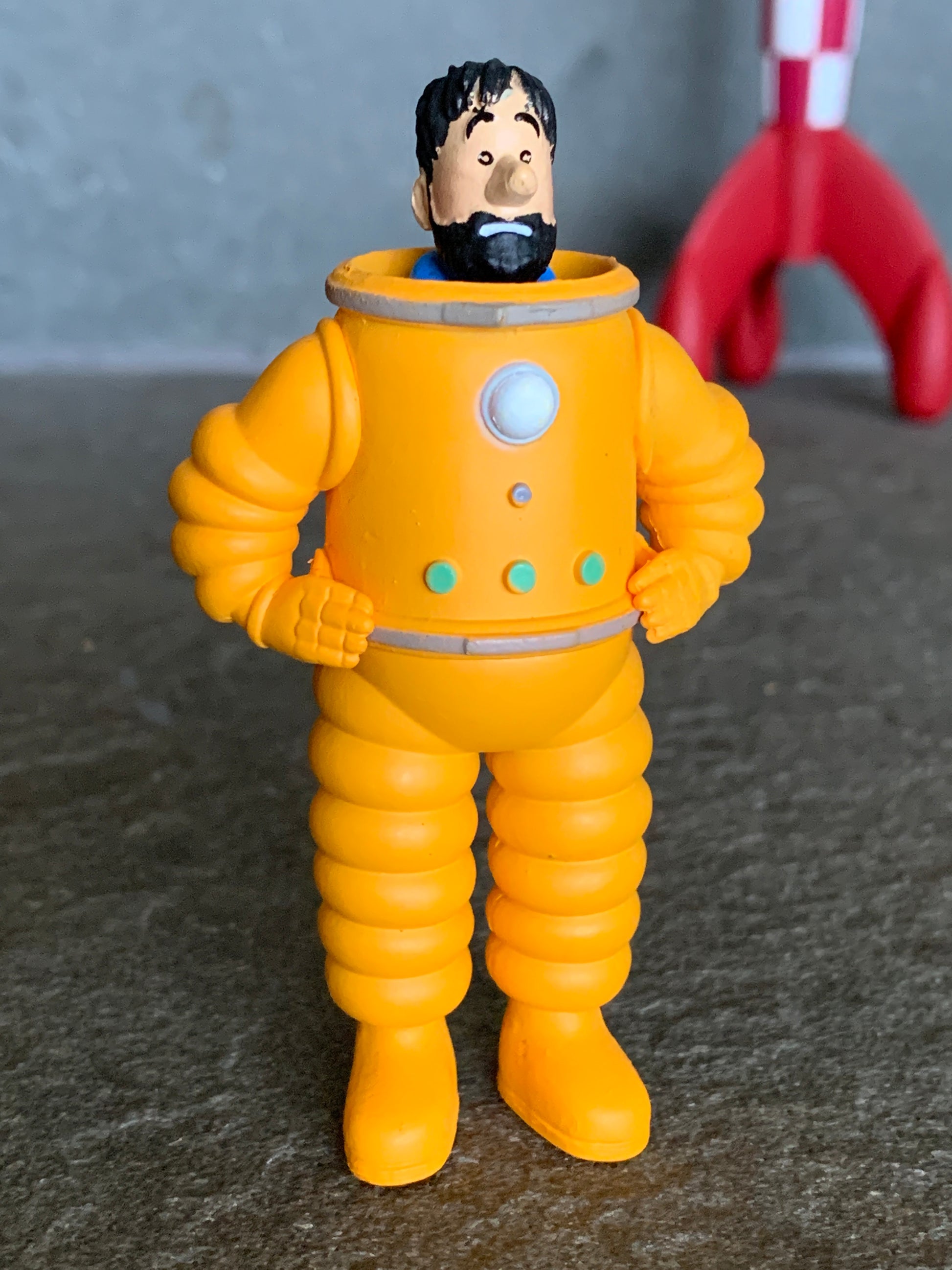 Captain Haddock cosmonaut mini figure in an orange space suit with hands on his hips, standing in front of a red and white checkered Tintin moon rocket.