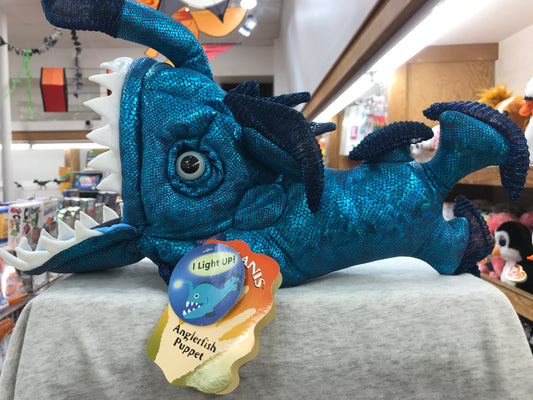 Folkmanis Anglerfish Puppet with LED lure, movable mouth and eyes, displayed in toy store. Blue-scaled plush fish with glowing tag.