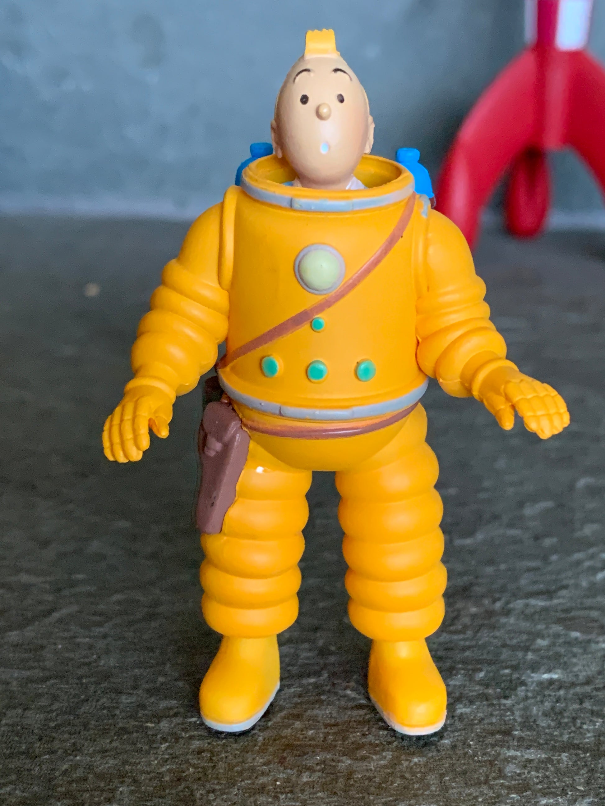 Close-up front view of the Tintin cosmonaut mini figure wearing a yellow spacesuit with a brown holster and a surprised expression, with a blurred red moon rocket in the background.