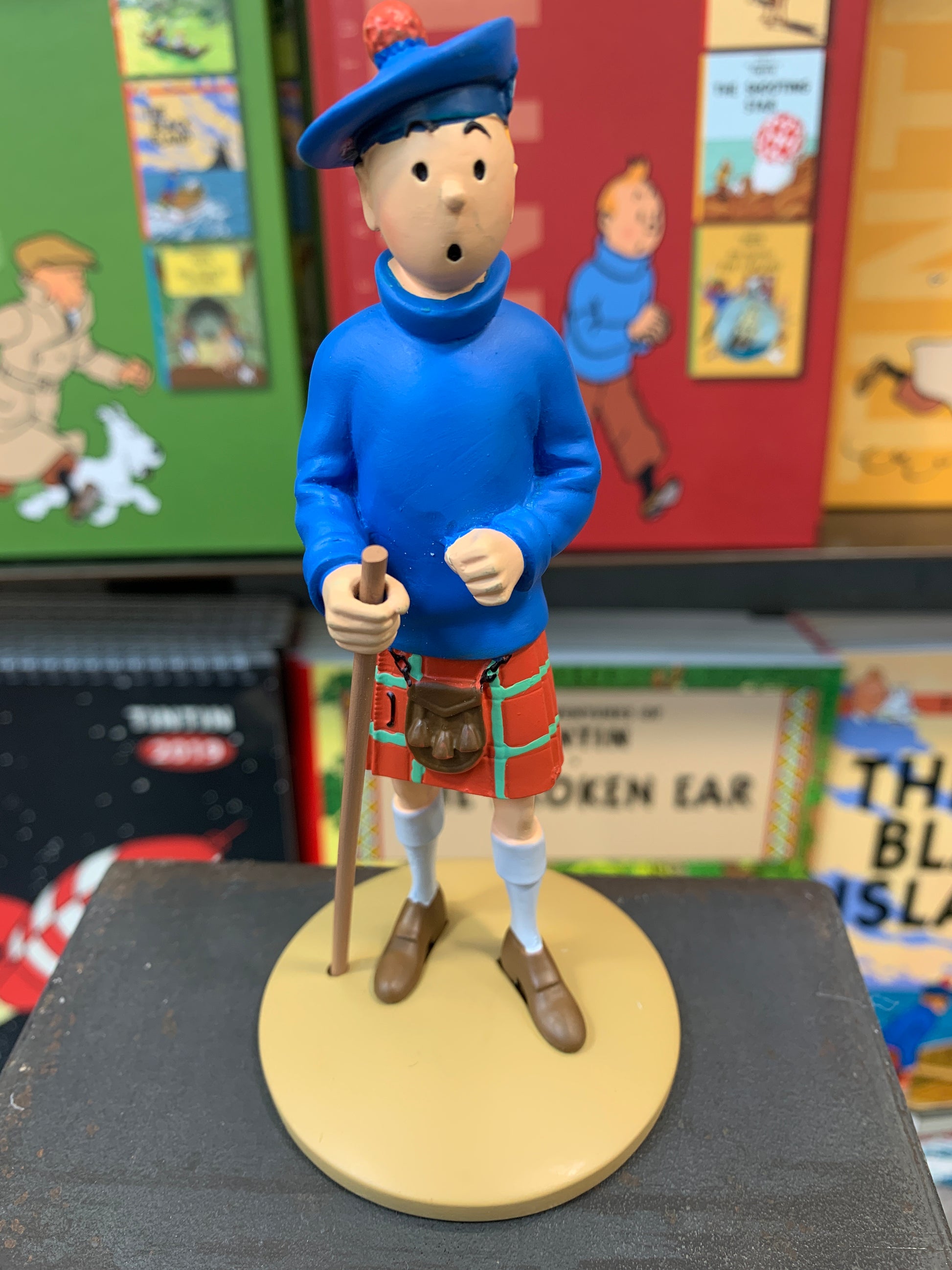 Tintin resin figure in Scottish kilt and blue top, holding walking stick, from The Black Island, displayed with comic books