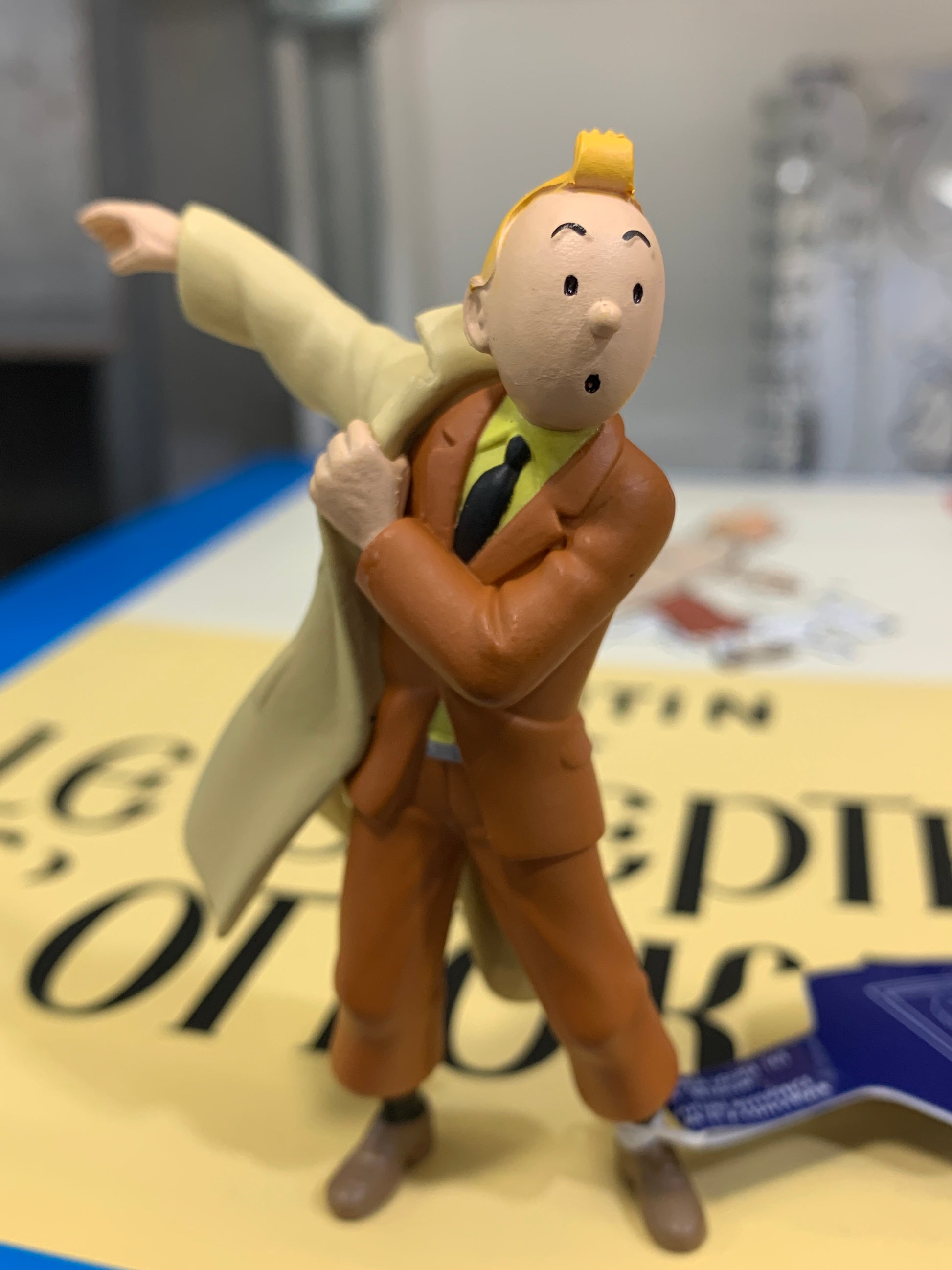 Tintin mini figure in a classic brown suit and tie, captured mid-motion as he puts on his iconic beige trench coat, standing in front of a blurred background.