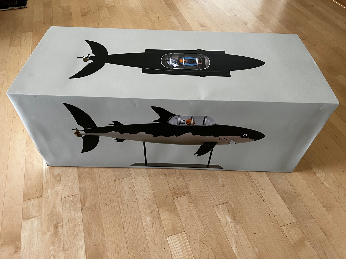 Large rectangular white packaging box for the Tintin 77cm Shark Submarine model from Red Rackham's Treasure. The top and side of the box feature a large black graphic silhouette illustration of Professor Calculus's shark-proof submarine, showing Tintin and Snowy visible inside the cockpit canopy. The box is resting on a light-colored hardwood floor.